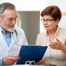 Stock photo of mature female patient wearing glasses discussing health complaints with doctor in a medical exam, representing participation with AIBL.